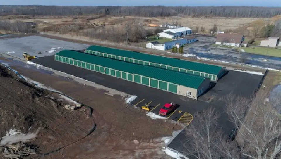 Aerial view of green-roofed storage units with cars parked on blacktop, near rural buildings.