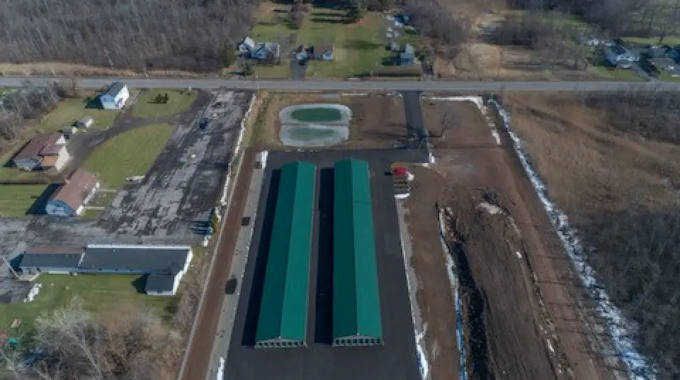 Aerial view of two long green-roofed buildings beside a road in a rural area with patches of snow.