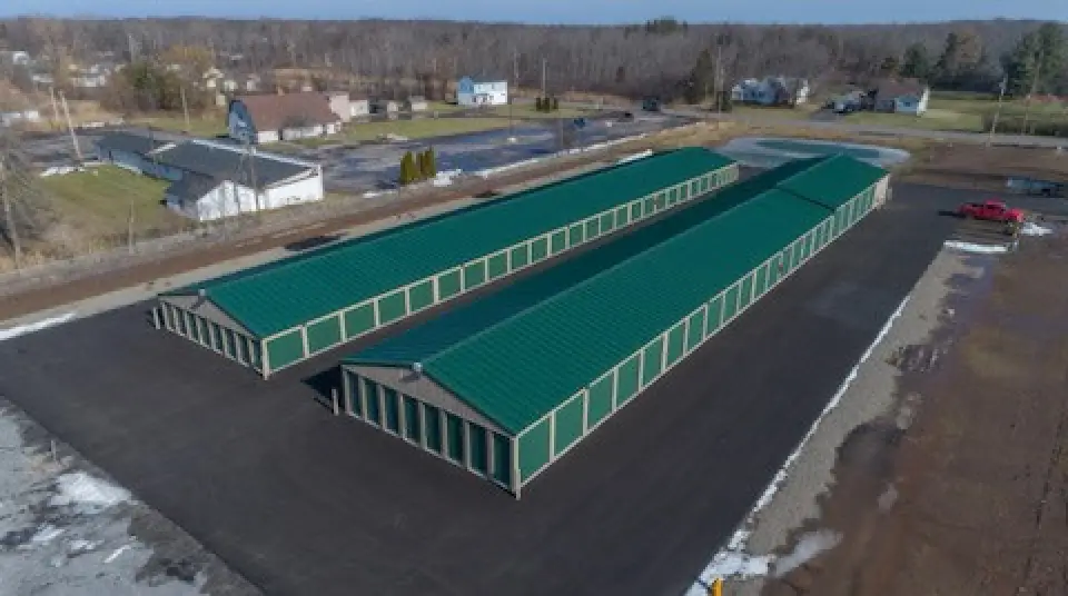 Aerial view of two long green-roofed storage buildings on a paved lot in a rural area.