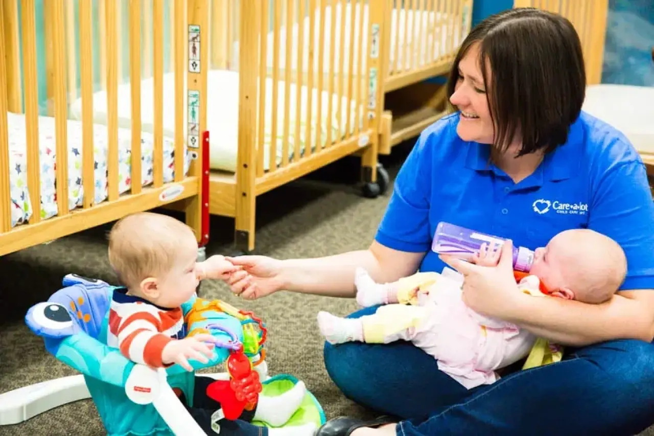 Smiling caregiver feeds a baby with a bottle while another baby sits and reaches for her hand.