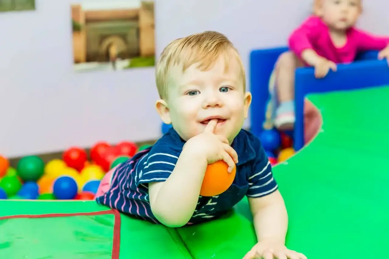 Smiling toddler lying on a green mat, holding an orange ball, with colorful balls behind.