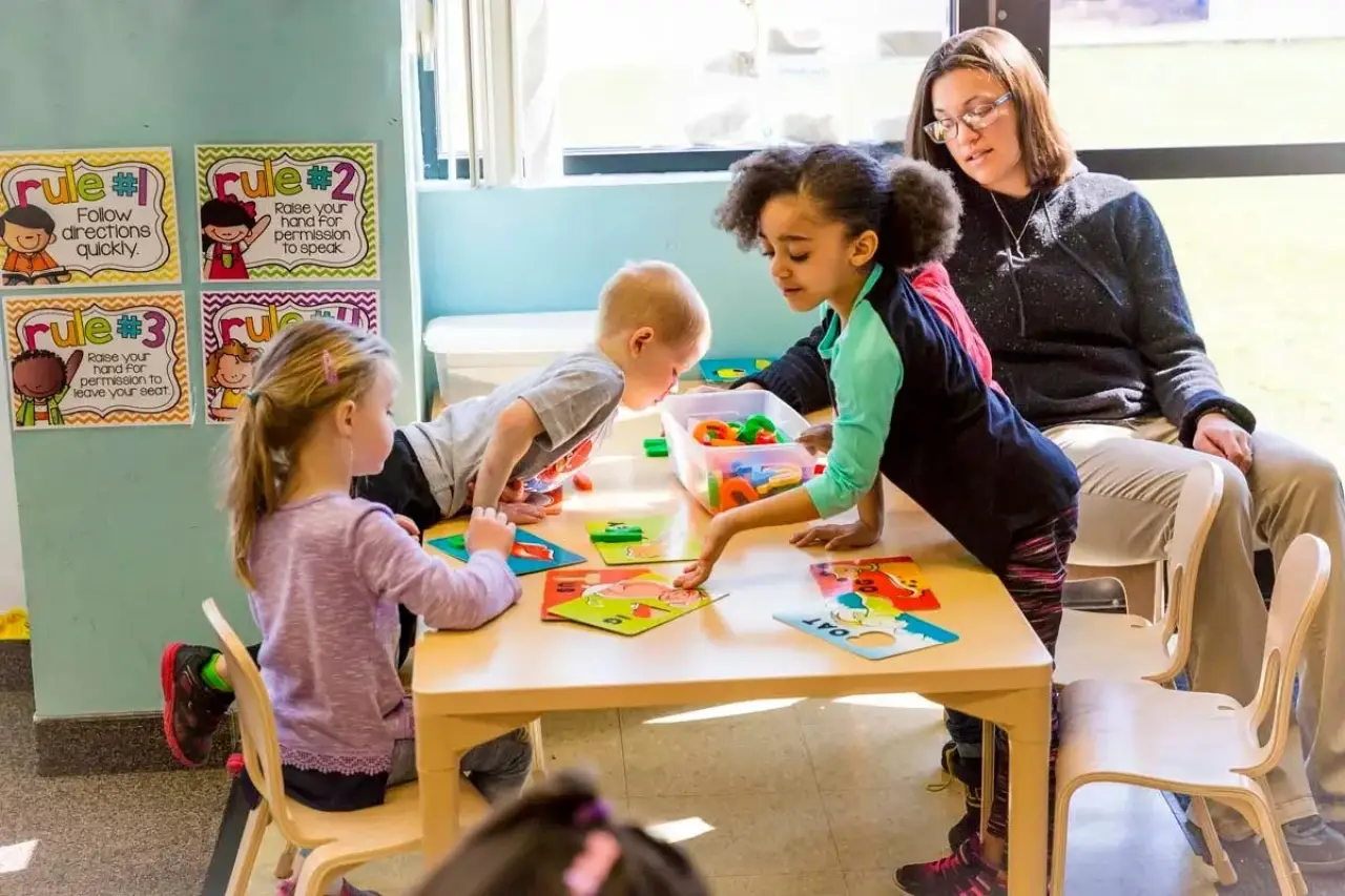 Four young children play at a table with puzzles while an adult supervises in a bright classroom.