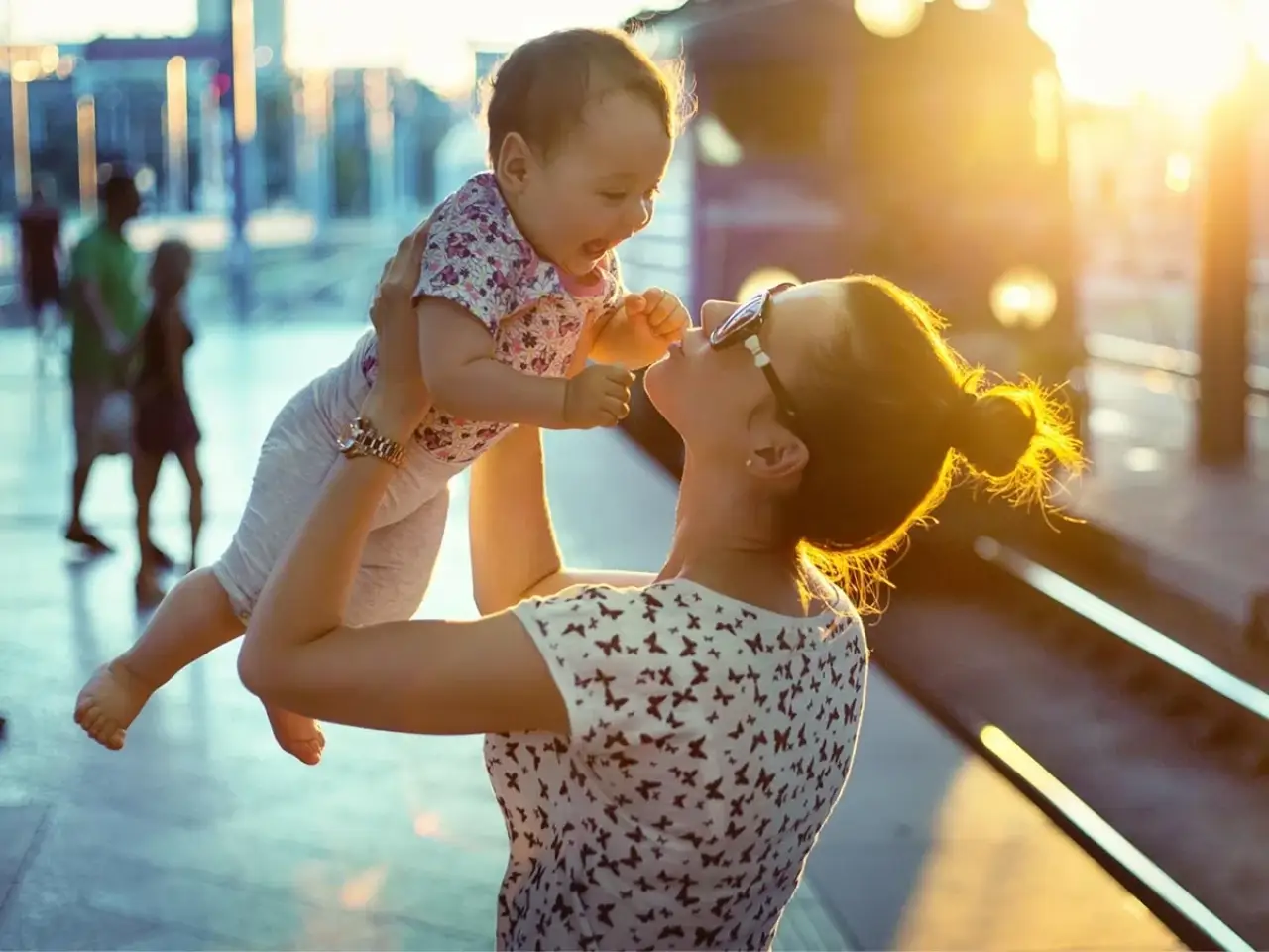 Woman holding up a smiling baby at a train station during sunset, both appearing joyful.