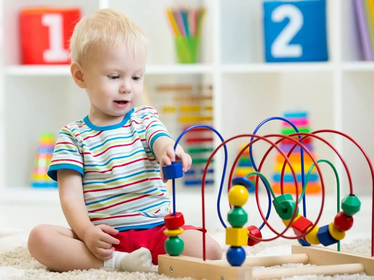 Toddler playing with a colorful bead maze toy in a bright, organized playroom.