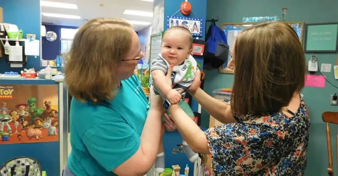 Two women smile while holding a happy baby in a colorful, toy-filled room.