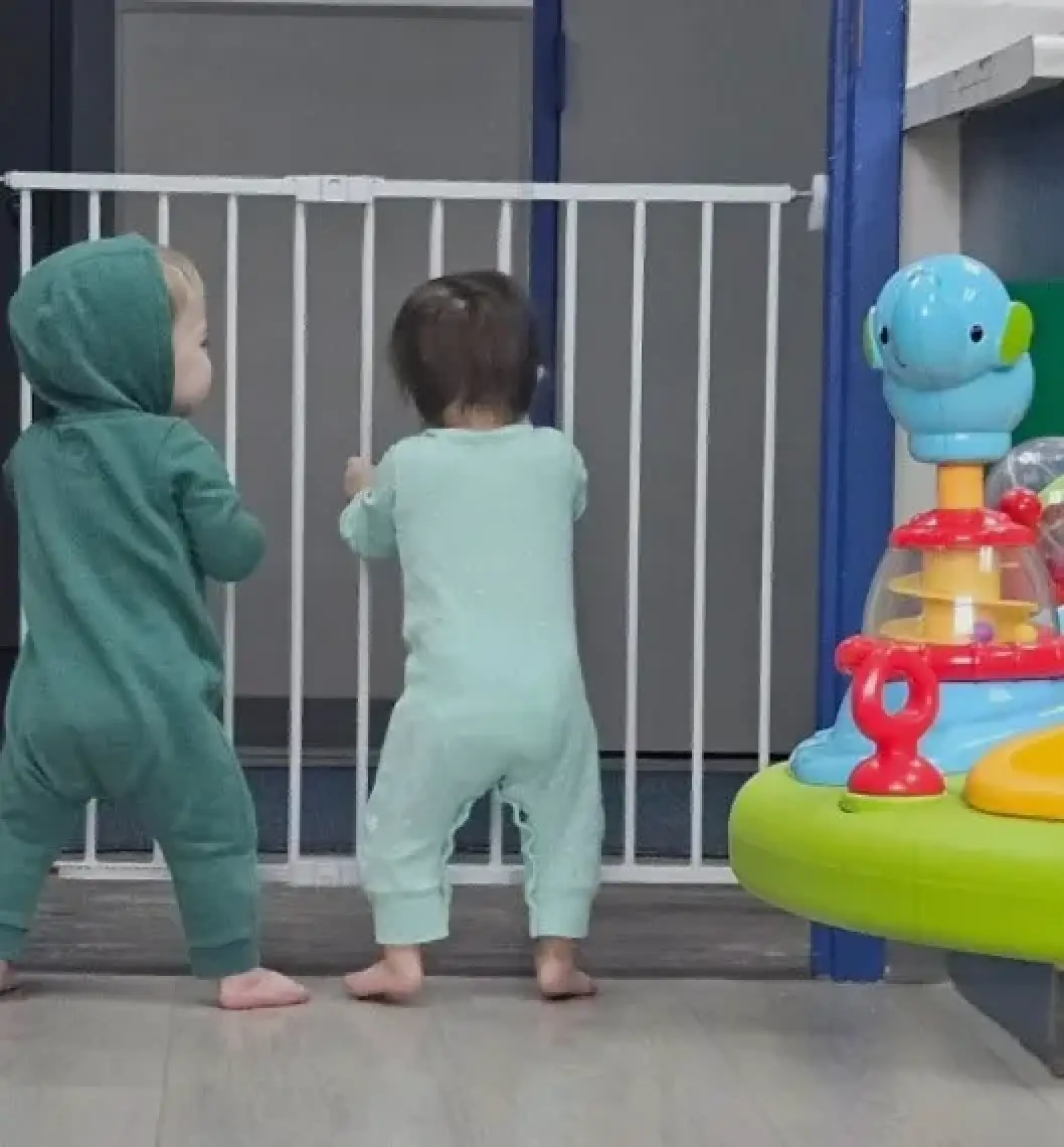 Two toddlers in pajamas stand by a white baby gate near a colorful toy in a playroom.