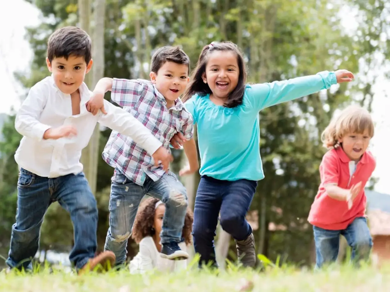 Four young children run and play outside on grass, smiling happily with trees in the background.