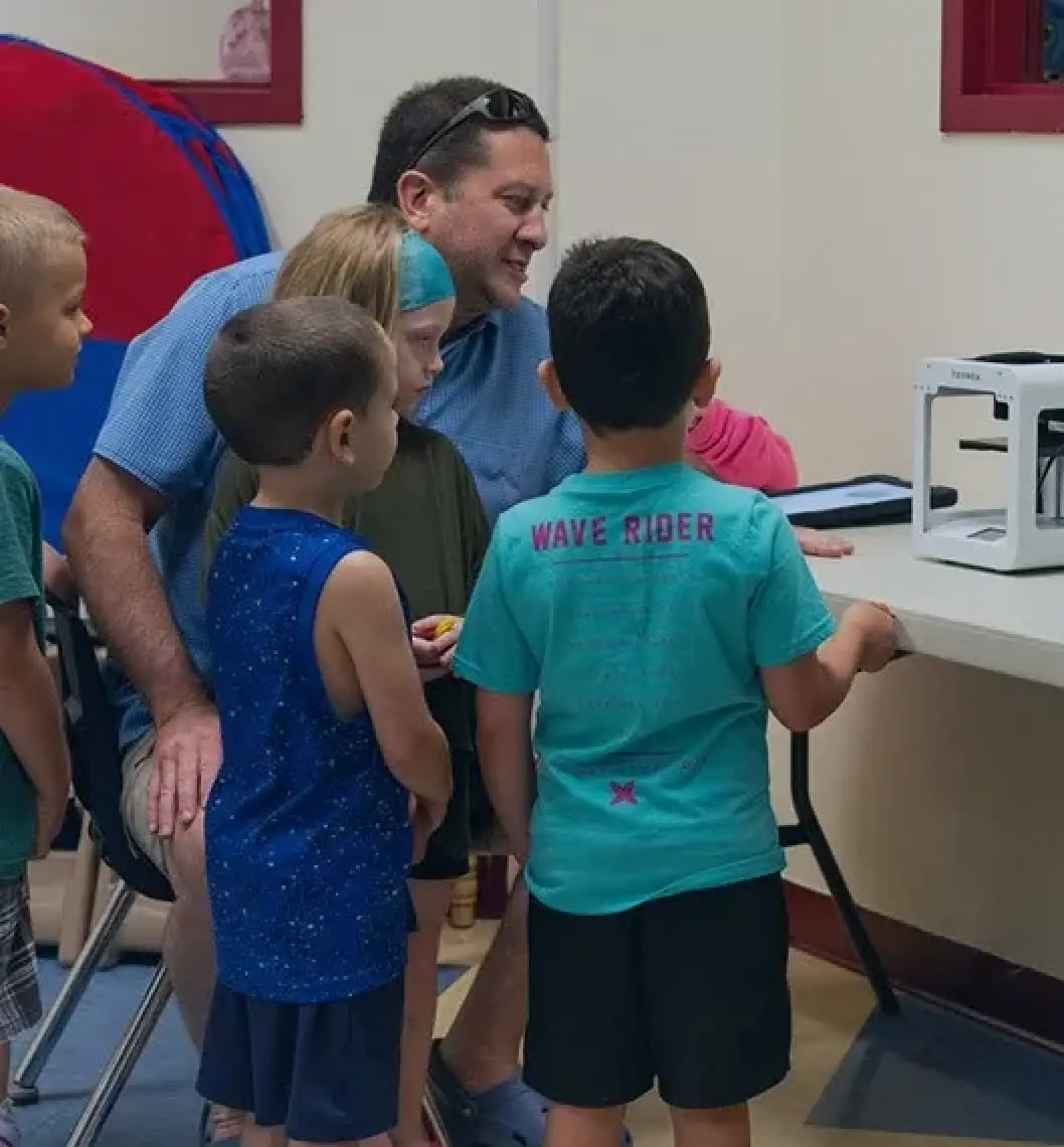 An adult and four kids watch a 3D printer at a table in a classroom setting.
