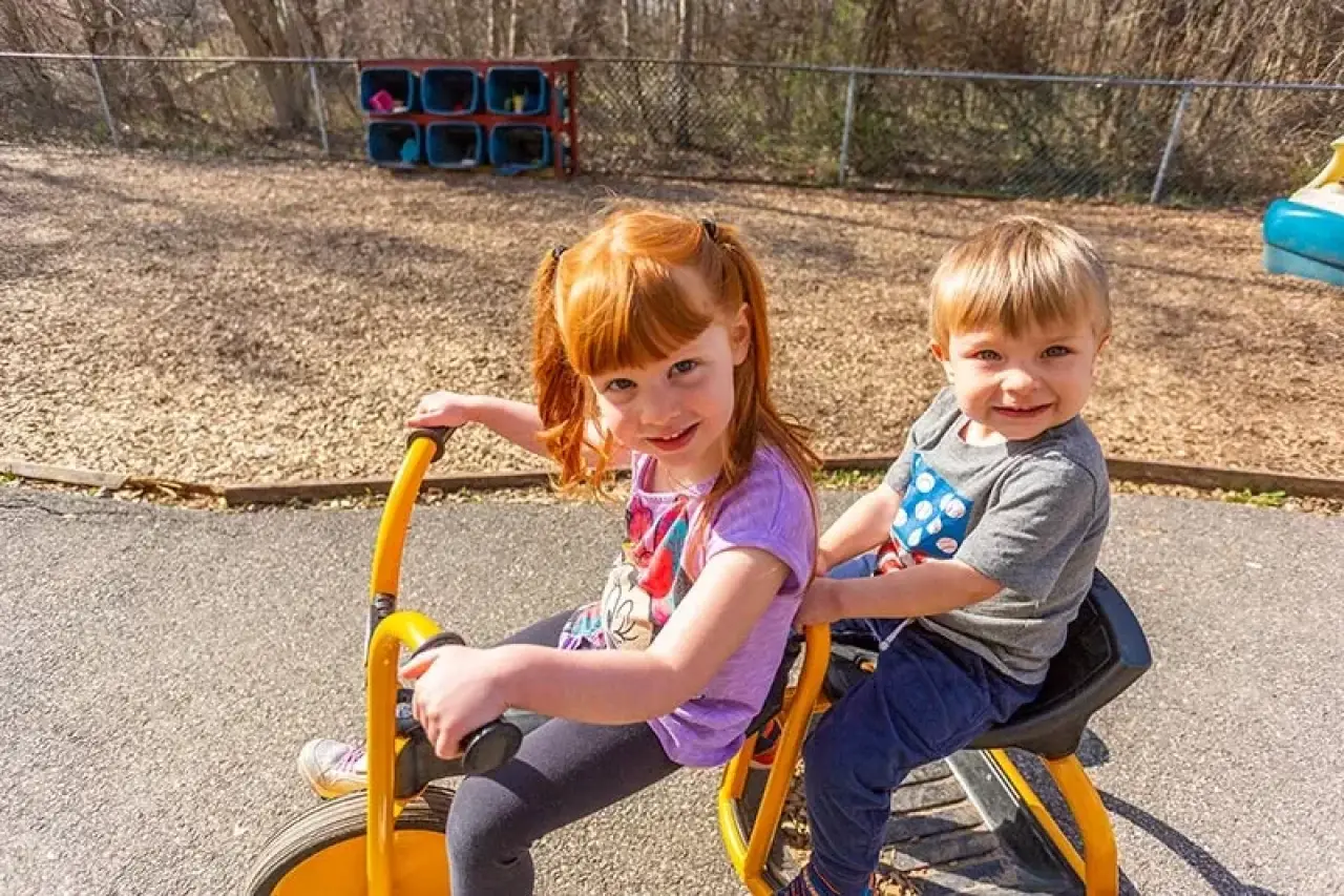 Two smiling children ride a yellow tricycle together on a sunny day at a playground.