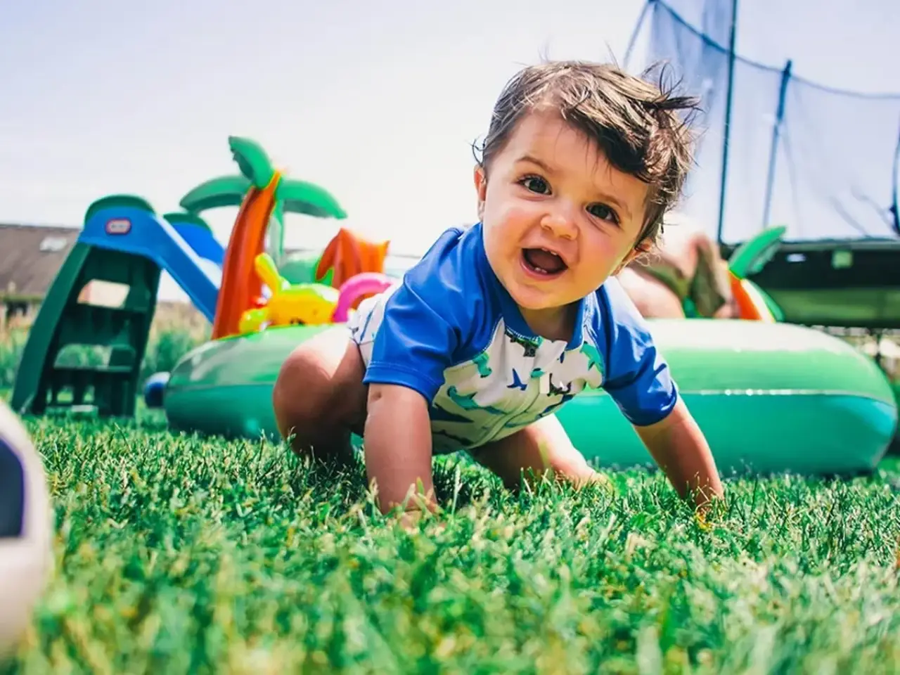A smiling baby crawls on green grass near an inflatable pool and outdoor toys on a sunny day.