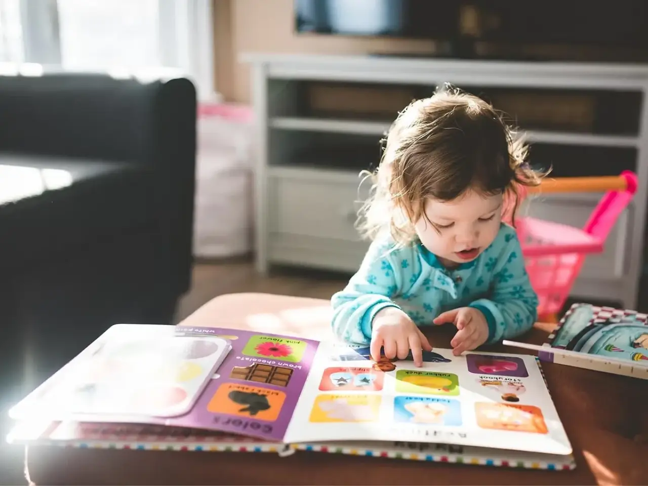 Young child in pajamas reads a colorful picture book at a table in a sunlit living room.