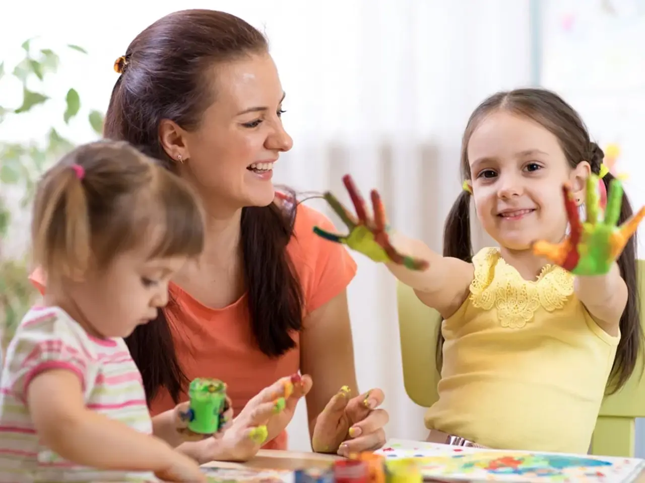 Woman and two young girls painting; one girl shows her colorful painted hands and smiles.