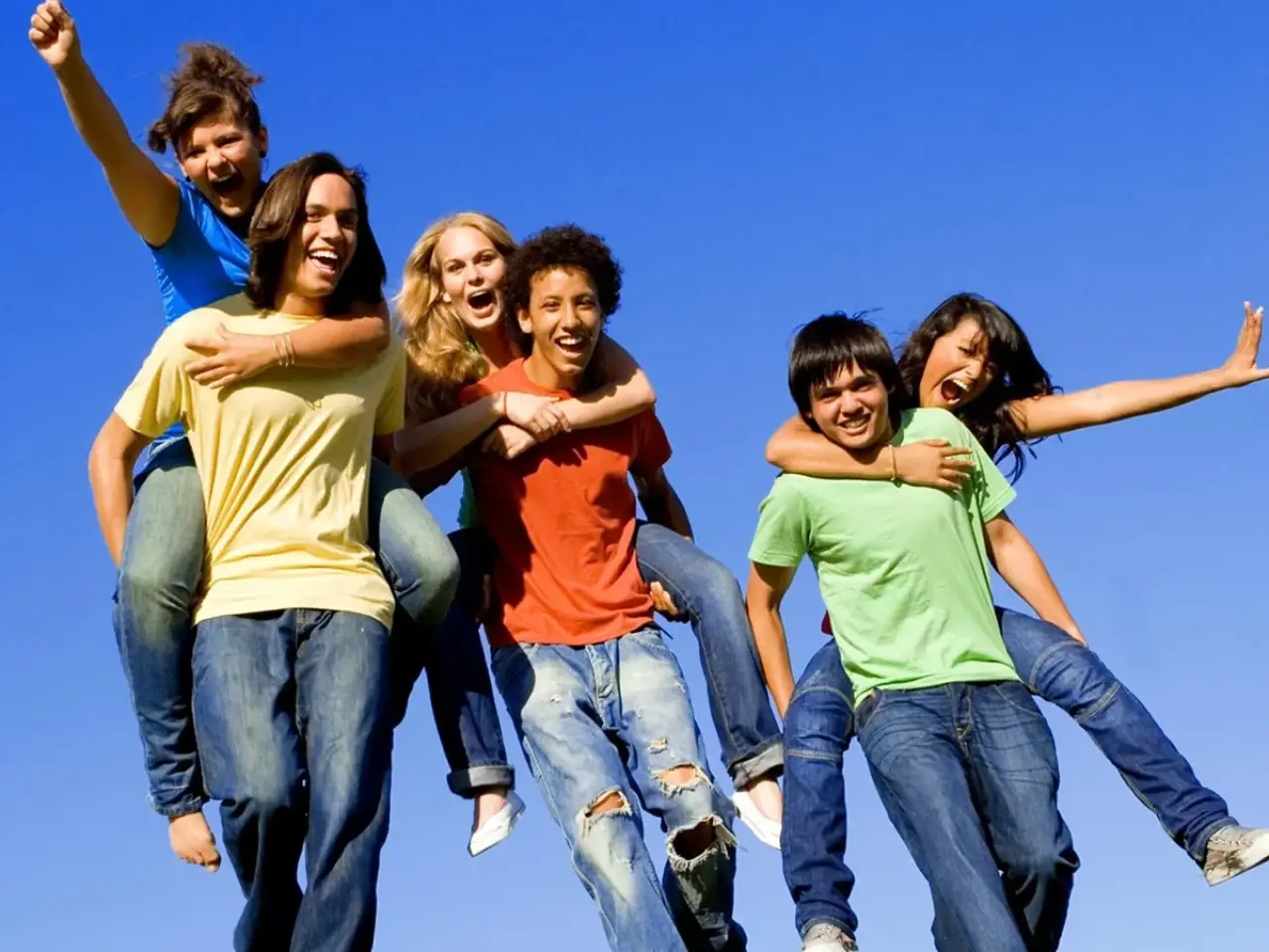 Six happy teenagers pose together outdoors against a bright blue sky, some giving piggyback rides.