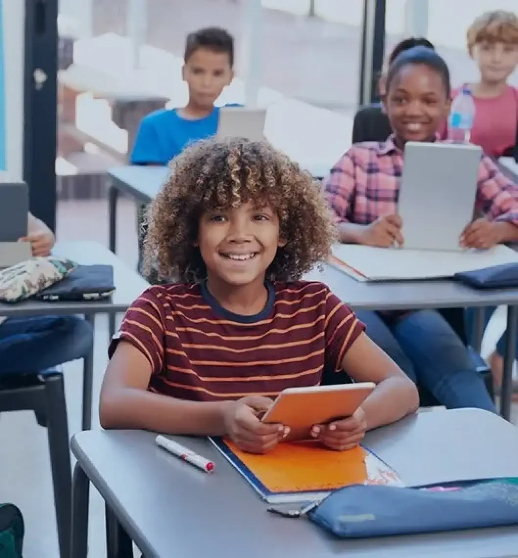 Smiling student holding a tablet, sitting at a desk in a classroom with other students.