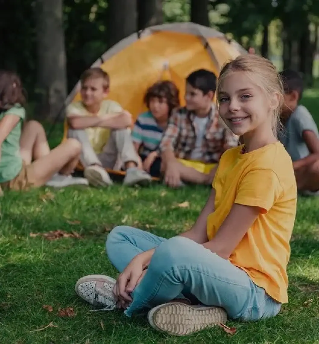 Smiling girl in yellow shirt sits on grass; other kids and a tent are in the background.