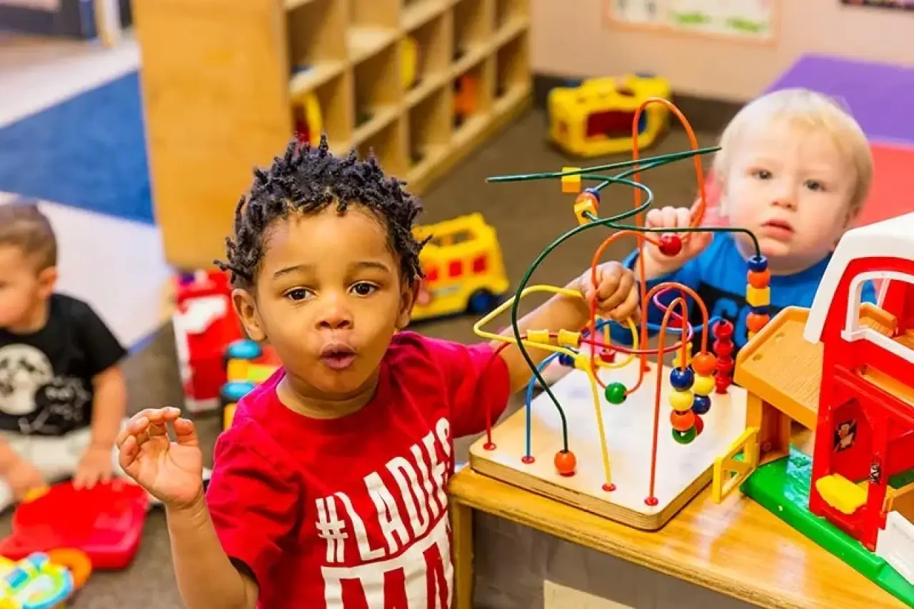 Two young children play with colorful toys at a table in a bright classroom or daycare.