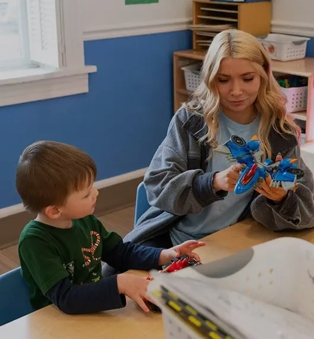 A woman and a young boy sit at a table, playing with toy vehicles in a classroom.