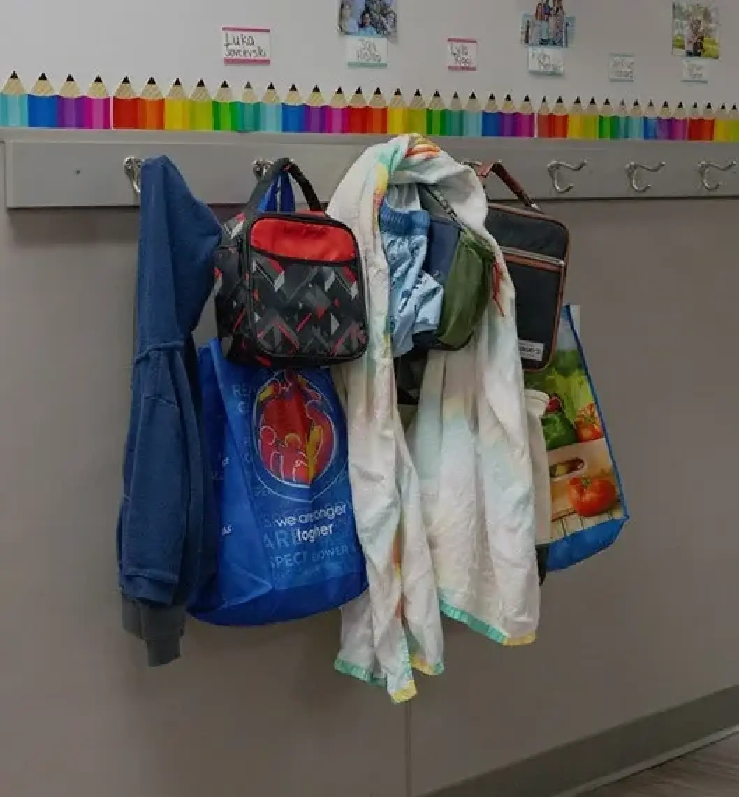 Childrens jackets, bags, and lunchboxes hang from hooks below a colorful border in a classroom.