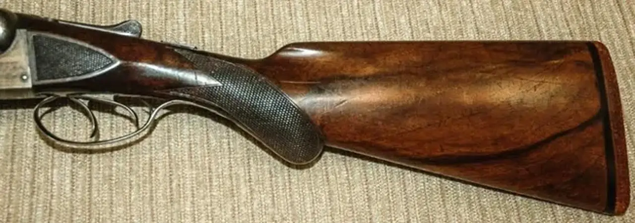 Close-up of a shotgun stock, showing wood grain and dark textured grip on a fabric surface.