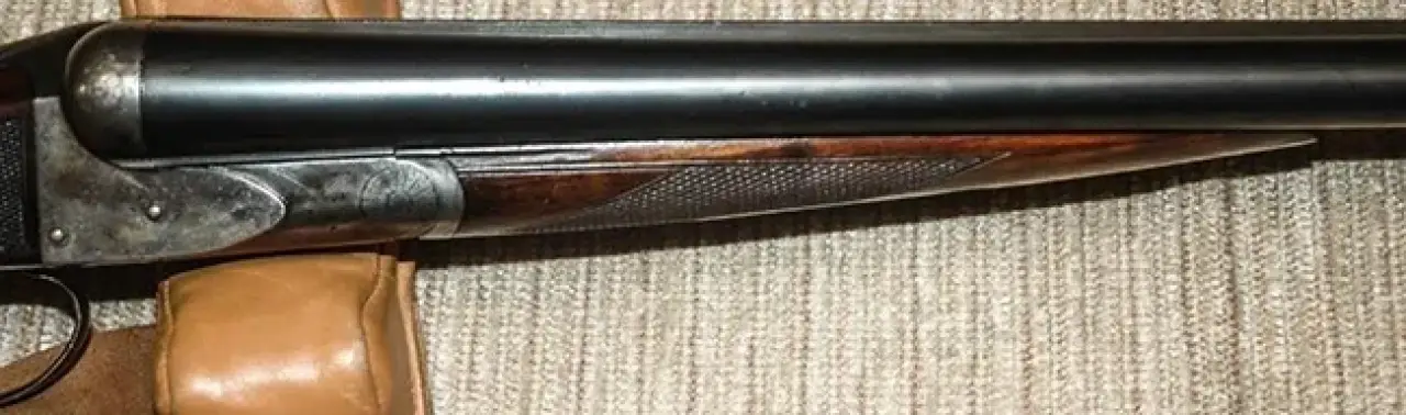 Close-up of a double-barrel shotgun resting on a beige surface, partially on a leather pad.