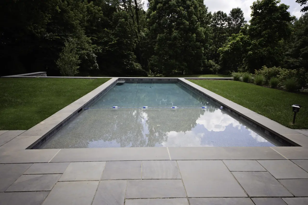 A rectangular swimming pool surrounded by grass and trees, with a stone patio in front.