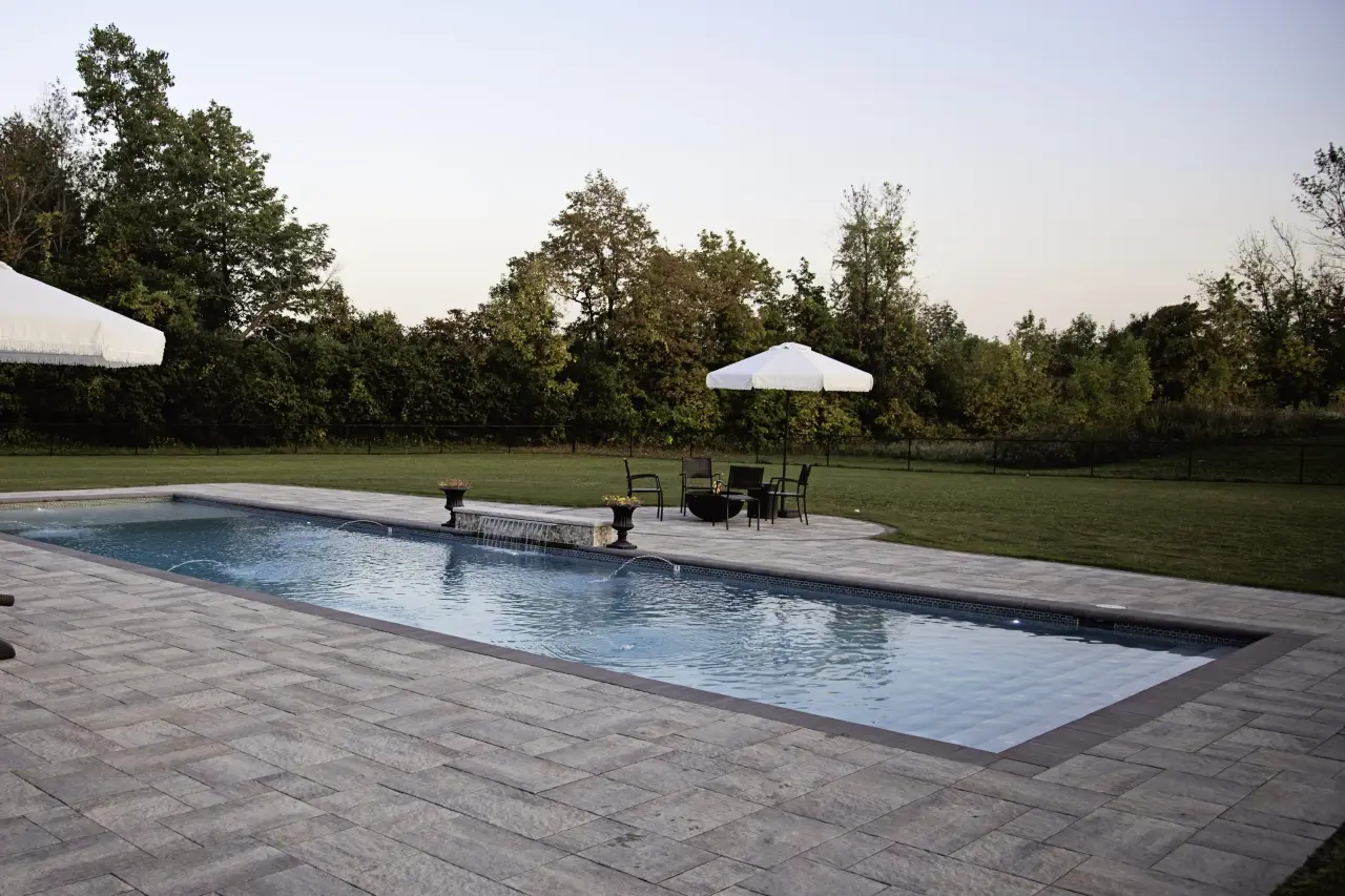 Rectangular backyard pool with patio, lounge chairs, umbrellas, and trees in the background.