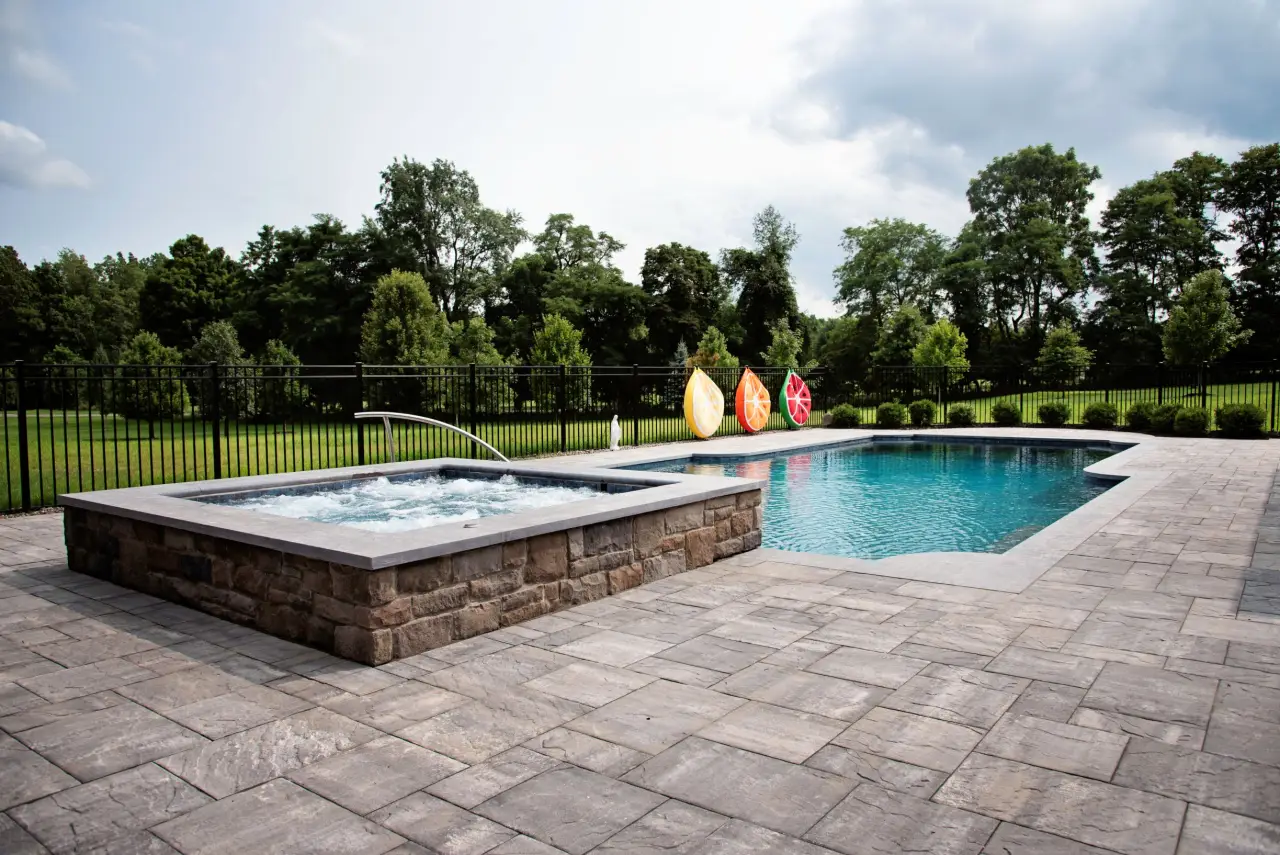 A backyard pool and hot tub with a stone patio, lawn, trees, and colorful kayaks by the fence.