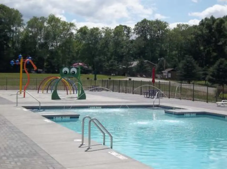 Outdoor pool with ladders, splash area, and playground sprays shaped like animals, trees in background.