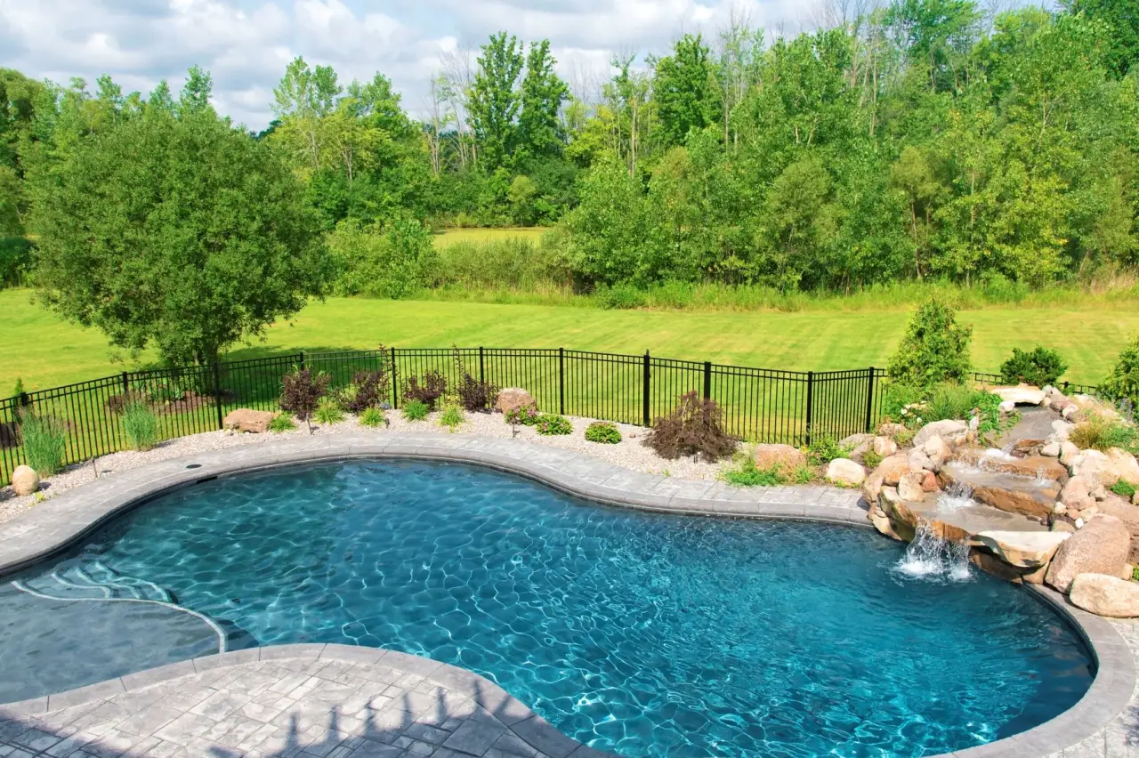 Backyard pool with a rock waterfall, stone deck, and a view of grassy yard with trees.