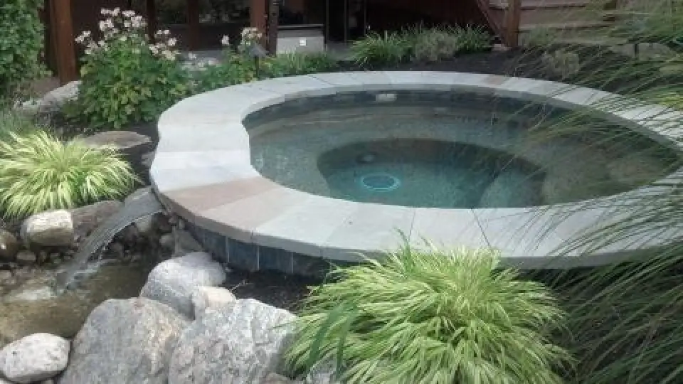 Round residential spa surrounded by ornamental grasses, flowers, and natural stone.