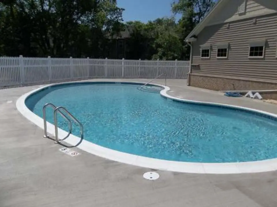 Curved outdoor swimming pool with metal ladders, near a beige house and white fence.