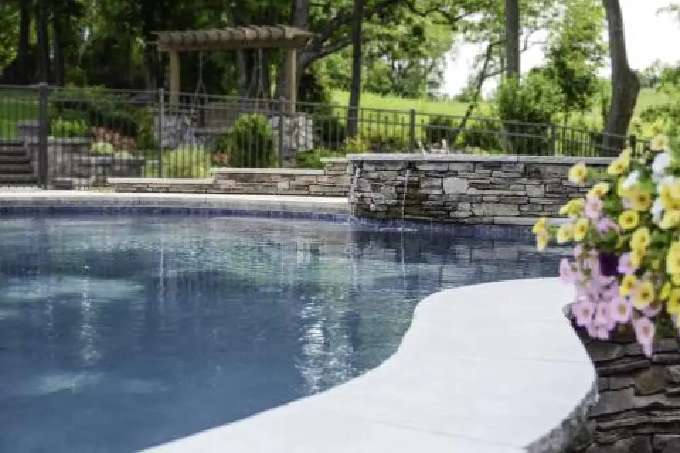 A curved pool with stone edging, flowers, and a small waterfall in a landscaped backyard.
