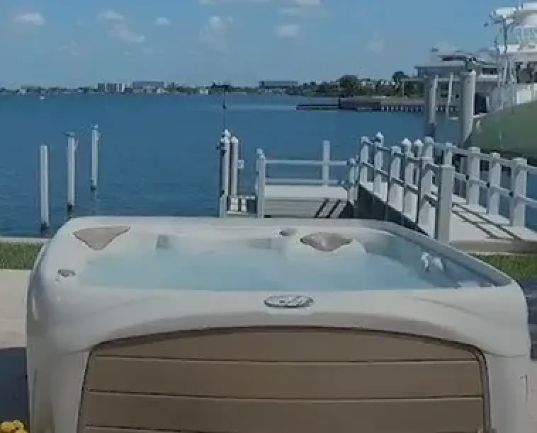A hot tub sits near a dock overlooking a calm bay with boats and buildings in the distance.