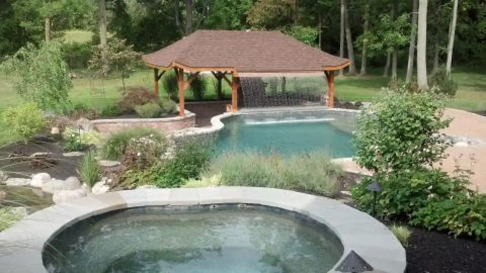 Backyard pool and raised spa surrounded by landscaping and a wooden pavilion.