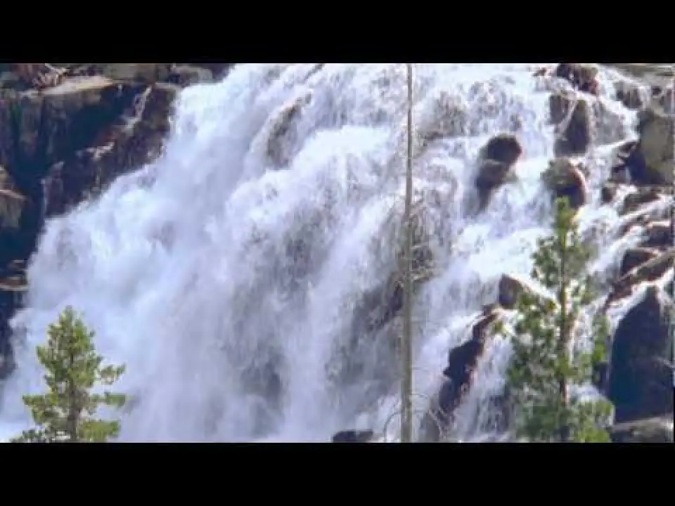 Waterfall rushing over rocky cliffs with evergreen trees nearby.