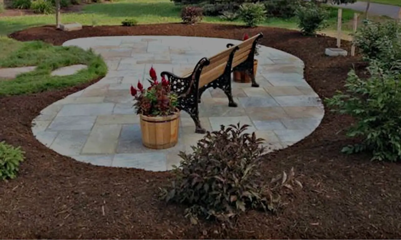 bean shaped stone patio surrounded by mulch with wodden bench on it
