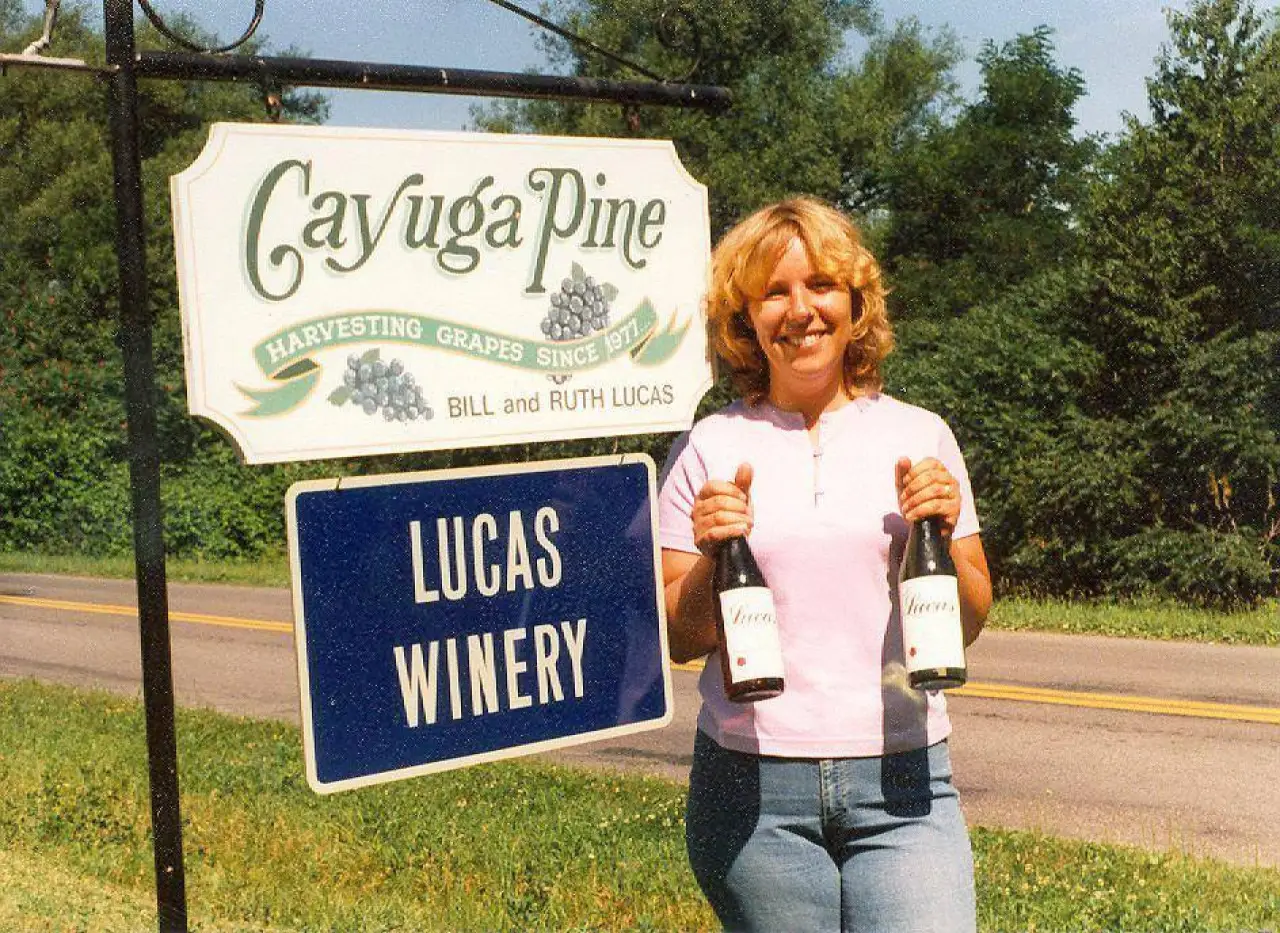 A woman holding two bottles beside the Cayuga Pine and Lucas Winery roadside sign