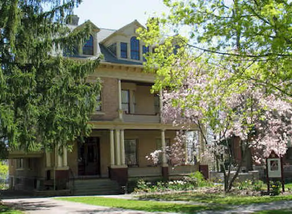 Large, older house with a porch, surrounded by green trees and blooming pink-flowered bushes.