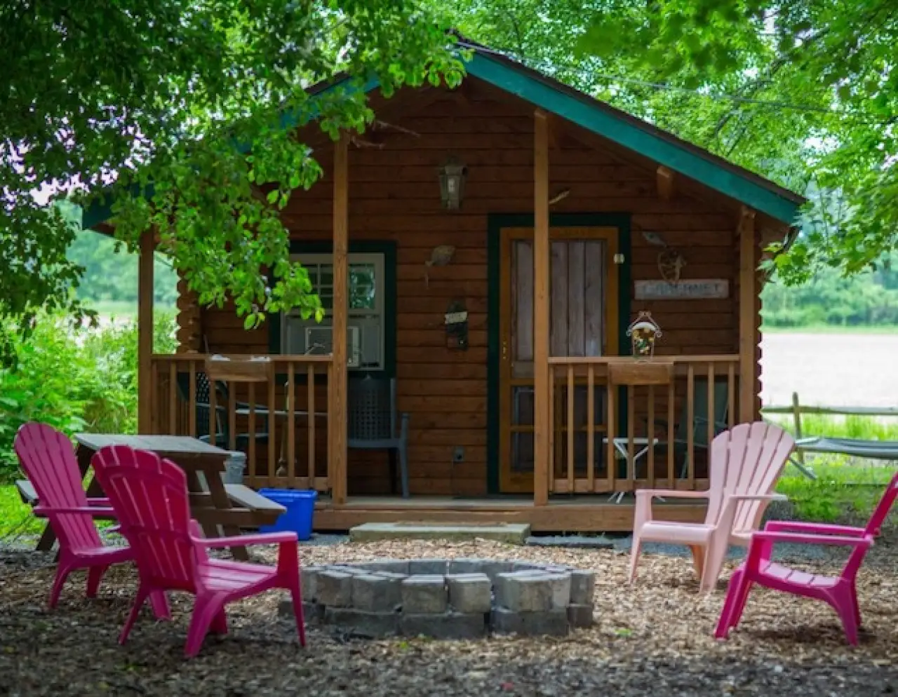 A cozy wooden cabin with a porch, surrounded by colorful chairs and a fire pit in front.