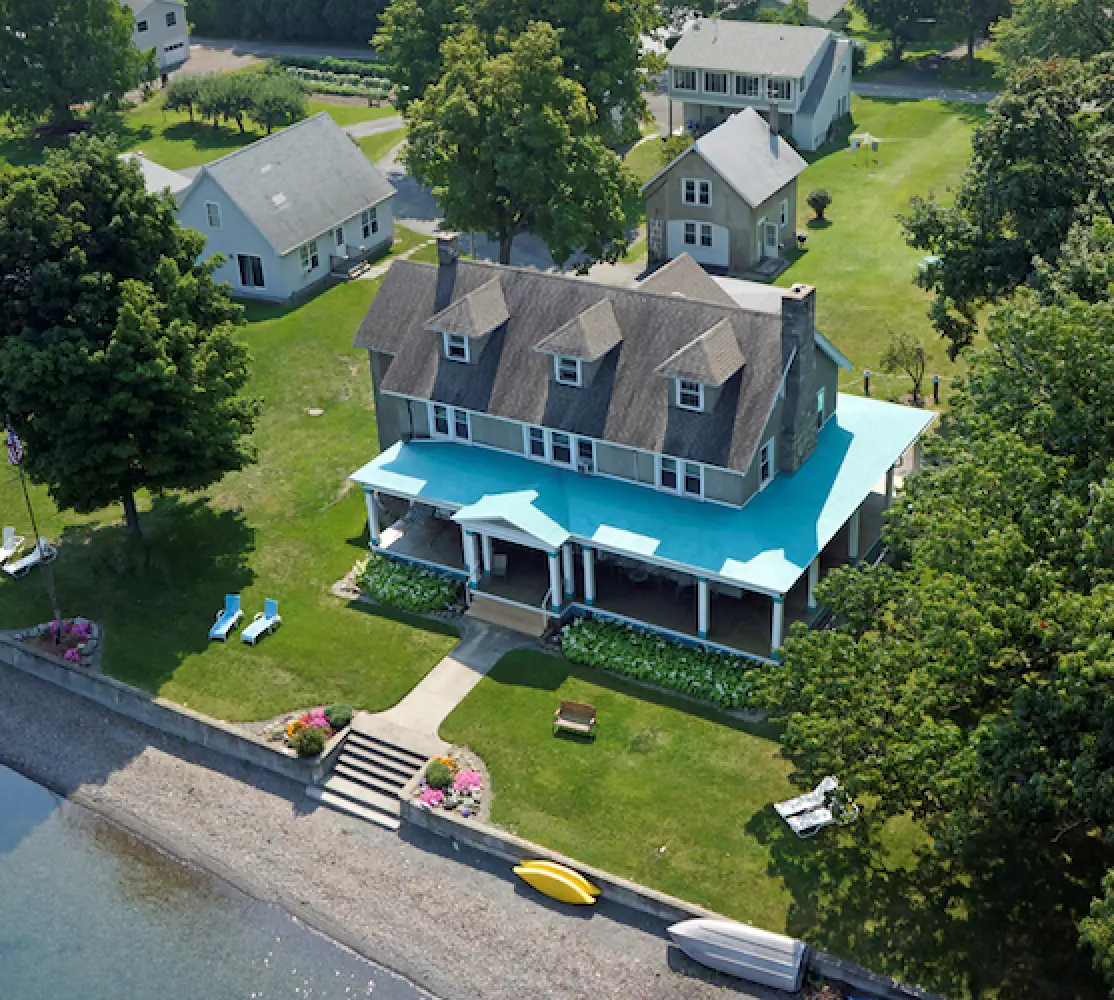 A large house with a bright blue porch roof sits by the water, surrounded by green lawns.