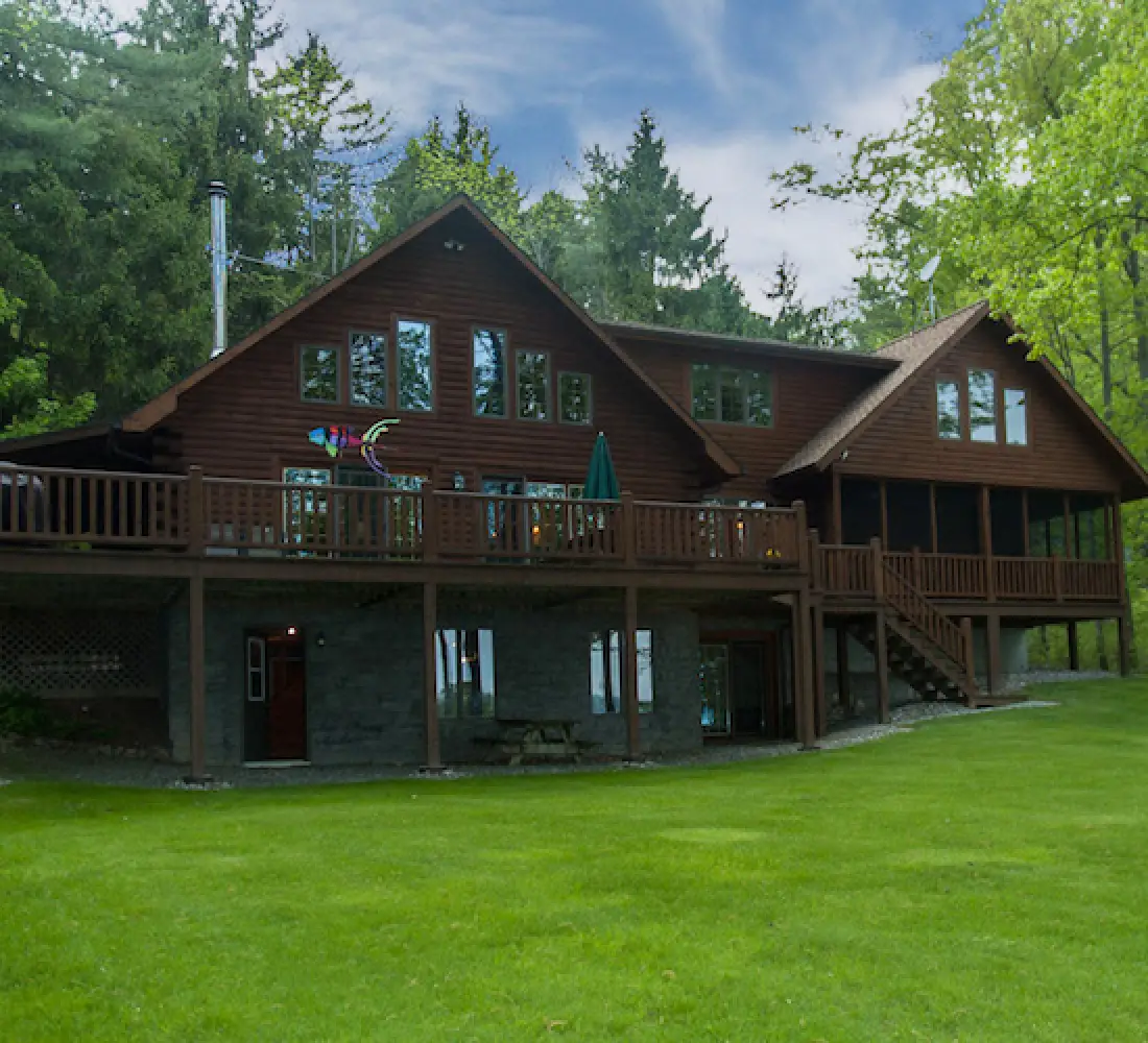 Large wooden cabin with a spacious deck, surrounded by green lawn and tall trees.