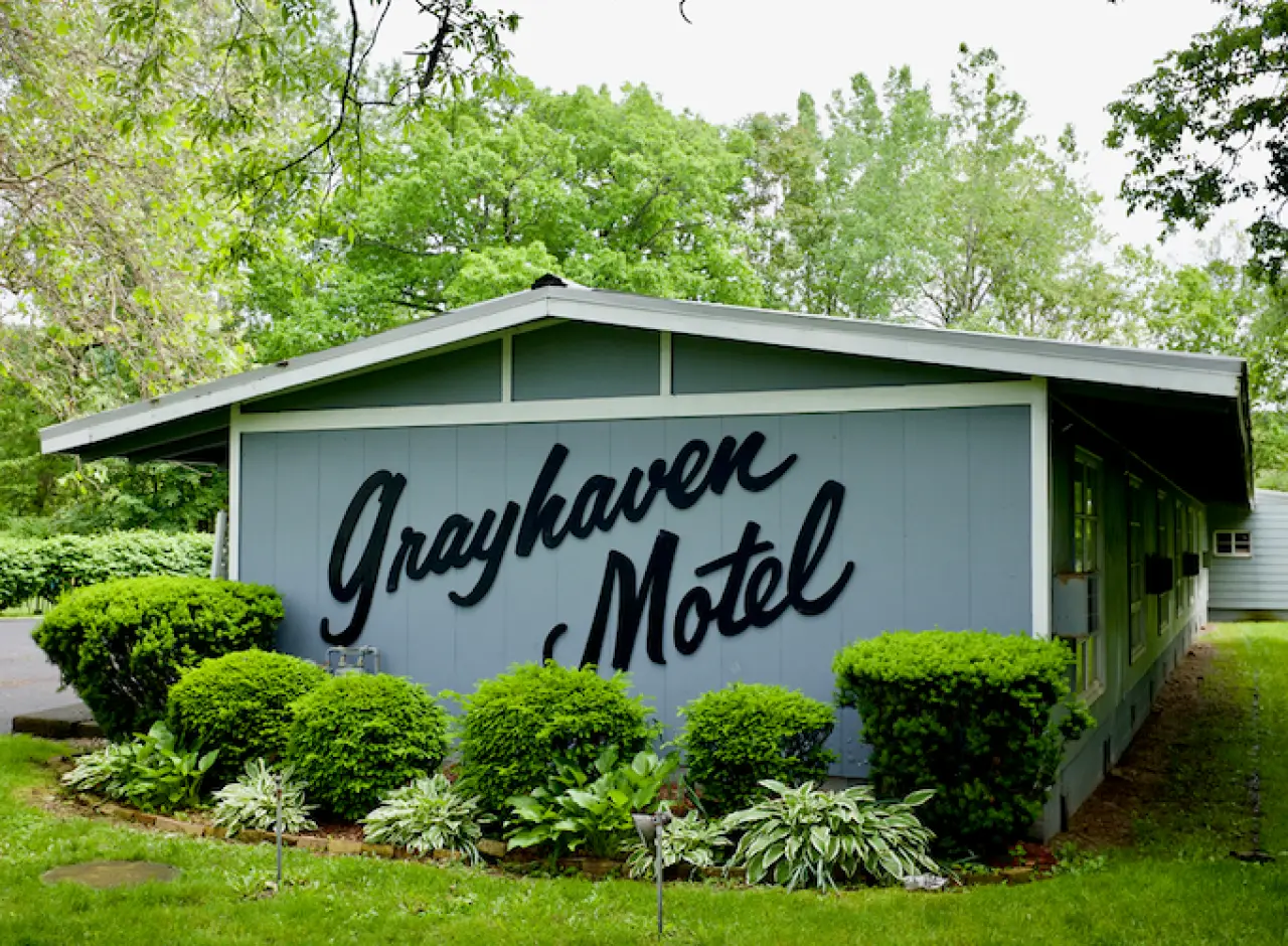 Grayhaven Motel sign on a blue building with green bushes and trees surrounding it.