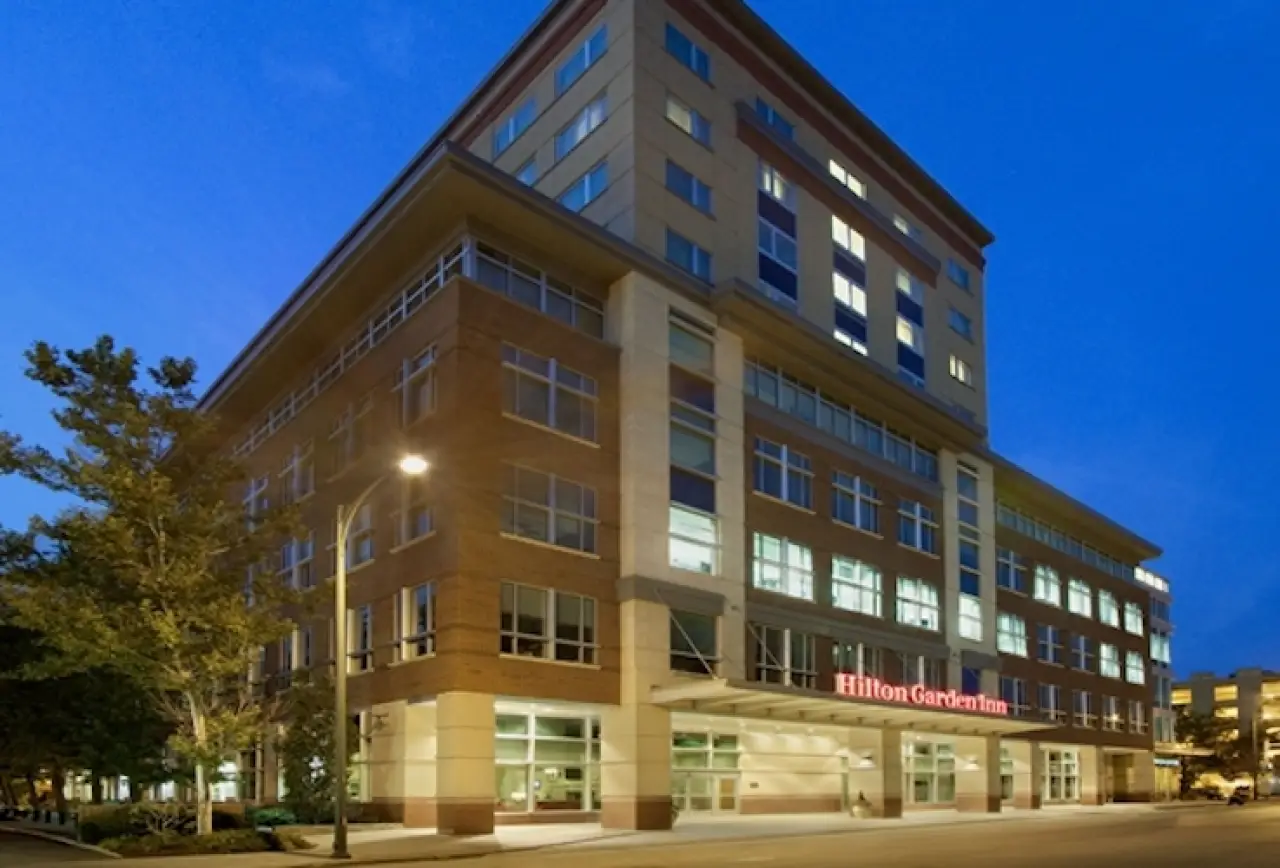 Modern Hilton Garden Inn hotel exterior at dusk, with lit windows and a tree nearby.
