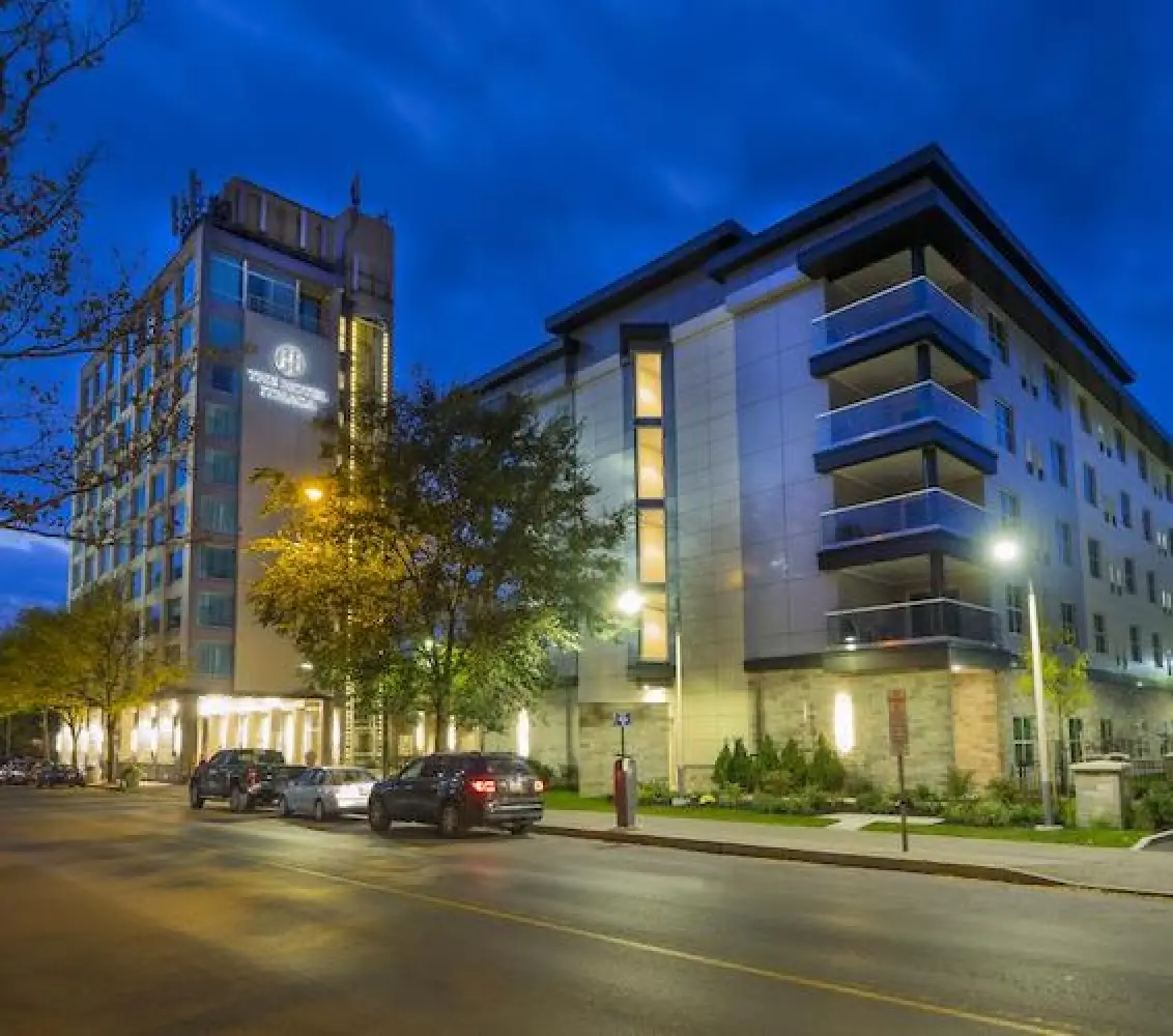 Modern hotel buildings with lit windows and parked cars along a city street at dusk.