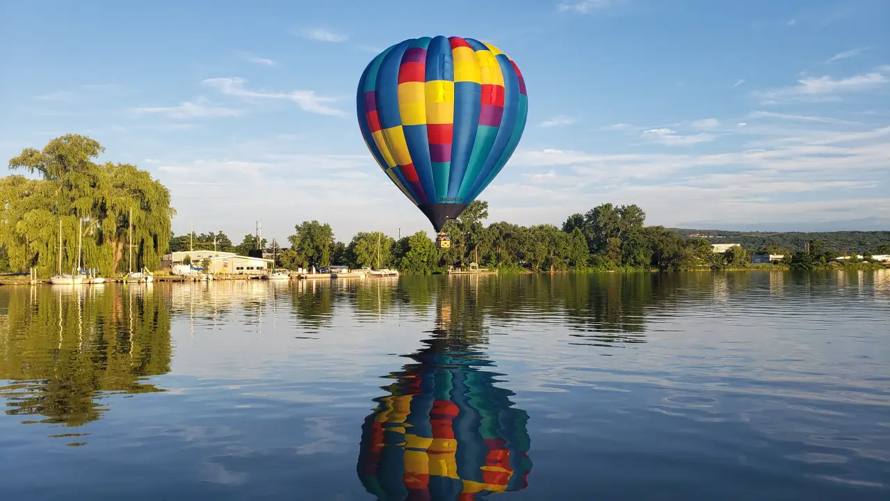 A colorful hot air balloon floats low over a calm lake, reflecting on the water’s surface.