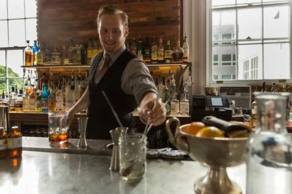 bartender smiling while mixing a drink behind a bar counter with various bottles and fruits