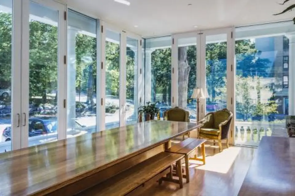 modern sunlit room with a long wooden table, chairs, and large windows overlooking trees