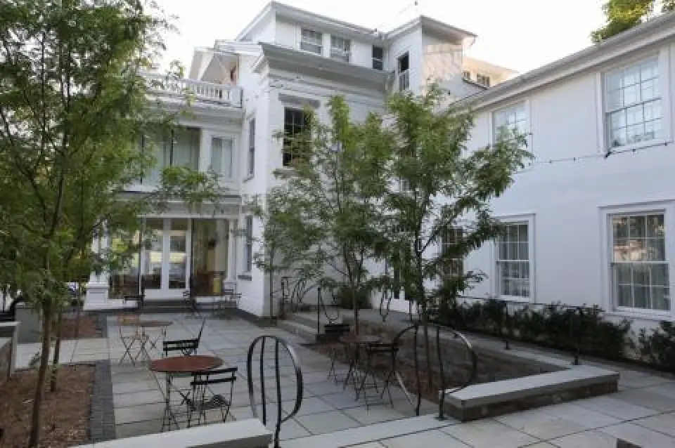 patio with tables and chairs surrounded by trees, adjacent to a large white building