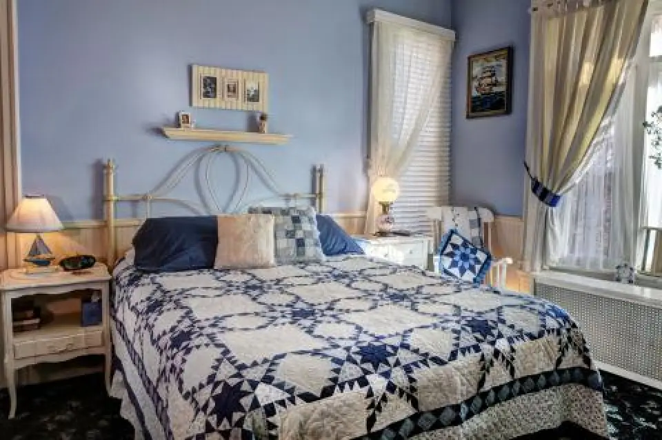 A cozy bedroom with a blue and white quilt, two nightstands, and sunlight streaming through curtains.