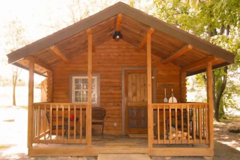 A small wooden cabin with a front porch, chairs, and trees surrounding it on a sunny day.